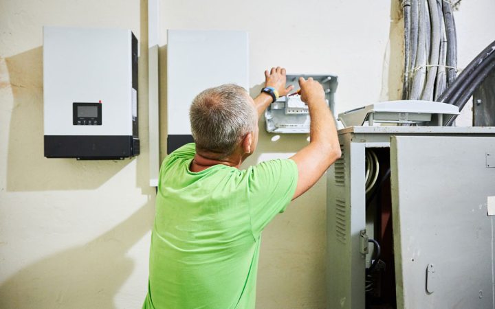 Electrician in bright green shirt installing or servicing wall-mounted solar inverters and electrical distribution equipment with exposed wiring and service panel