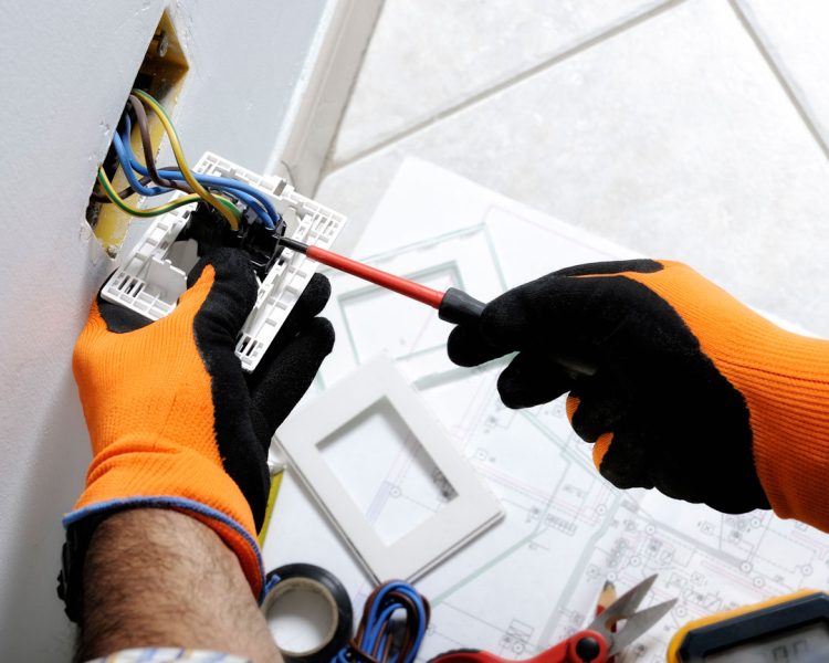 An electrician wearing orange and black safety gloves uses a screwdriver to connect wires to a new electrical receptacle, with wiring diagrams and tools laid out on the floor below.