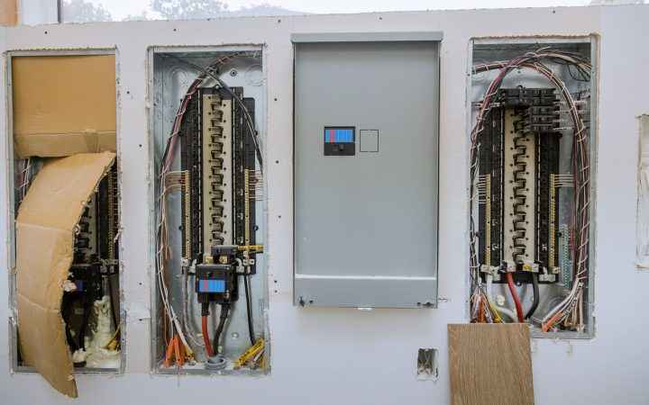 Wall-mounted electrical breaker panels during installation, showing exposed wiring and circuit breakers in open boxes alongside a closed gray metal panel cover.