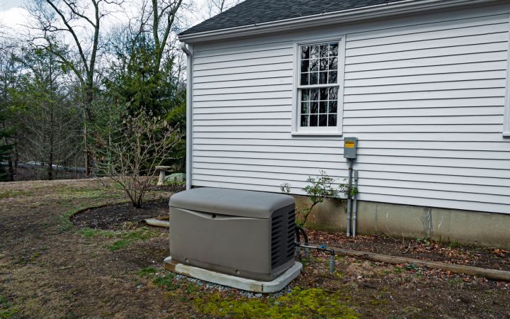 A whole-house generator installed beside a home with white siding, connected to an external utility box, set against a backdrop of trees and a lawn in early spring or late fall.